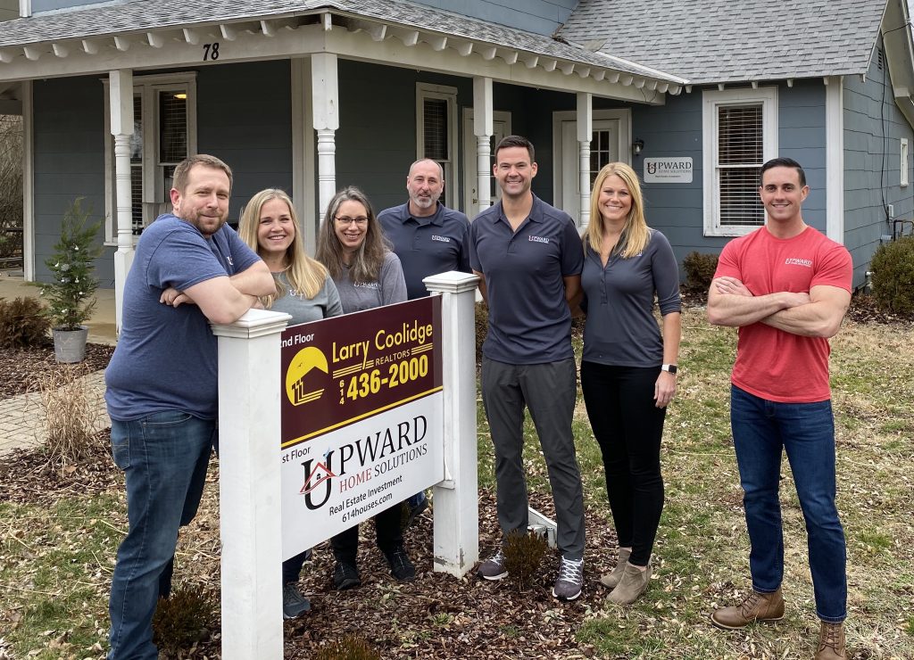 Seven men and women in front of a sign for the Powell real estate investment company Upward Home Solutions.