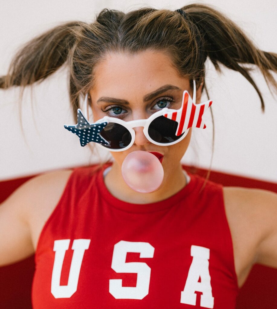 A woman wearing her brown hair in pig tails, a red USA tank top, and sunglasses blows a bubblegum bubble.