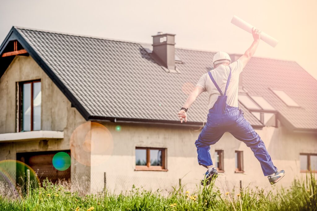 A man in blue overalls leaping in the air with a rolled up blueprint in his hand outside of a house.