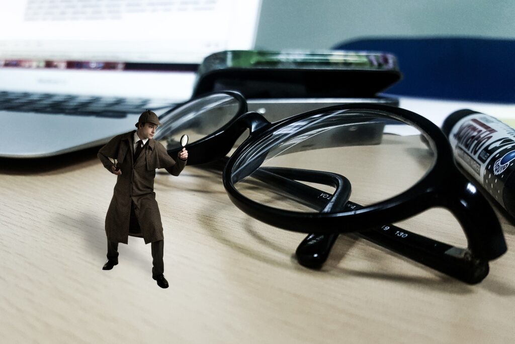 A miniature inspector holds a magnifying glass up to a pair of black eyeglasses.