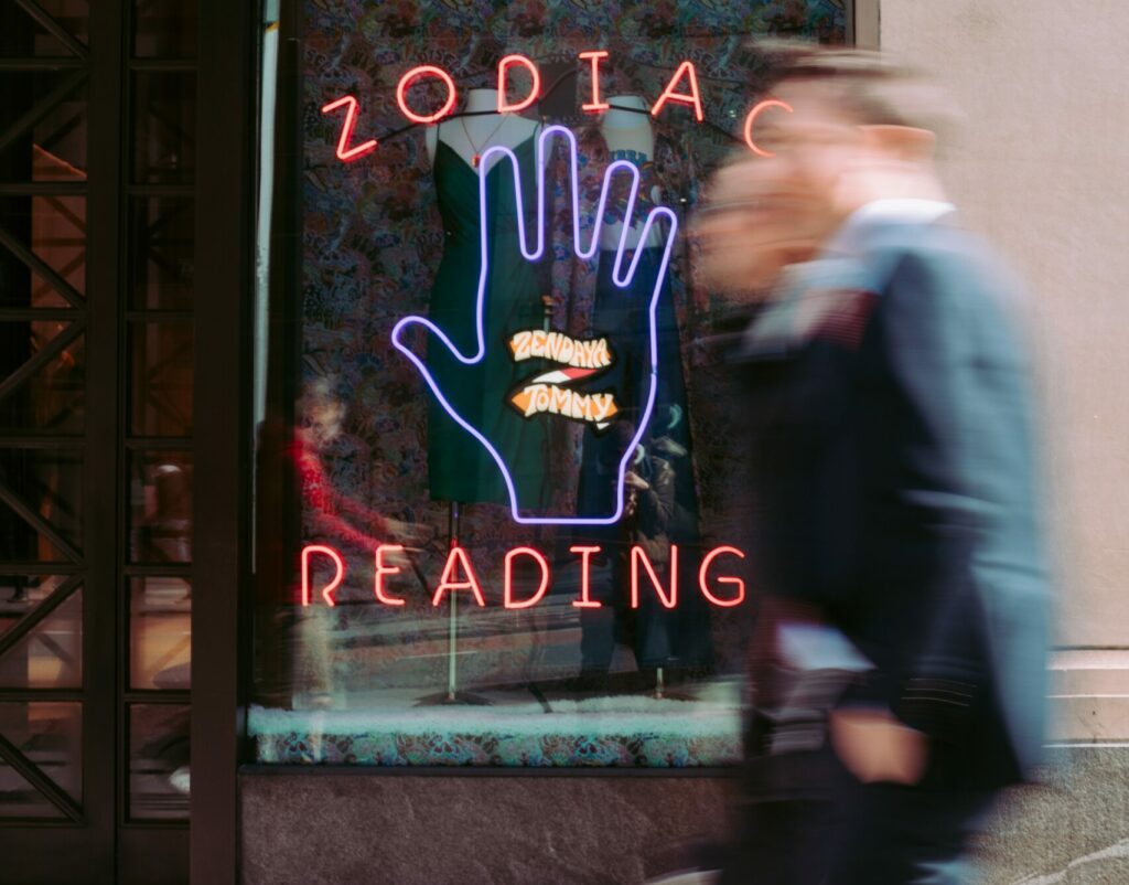 Two men walk past a shop window with a neon sign with the words "ZODIAC READING" around a neon purple hand.