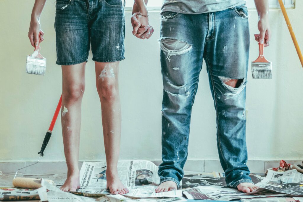 The bottom half of two house painters, holding hands, each with a paintbrush in their other.