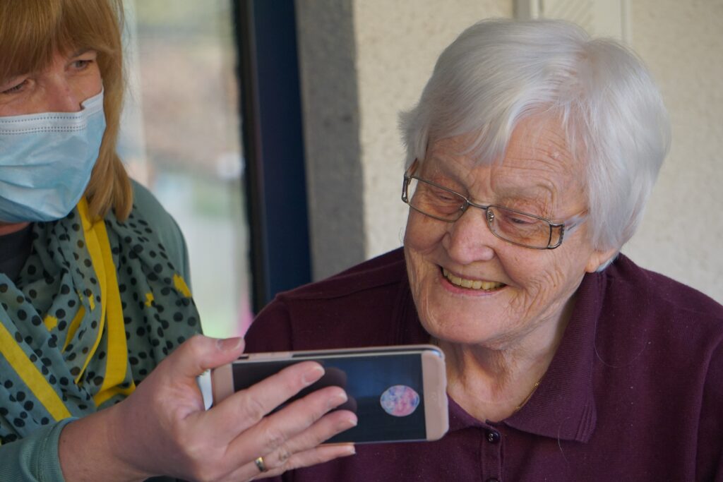 A senior woman smiles at a cell phone held in front of her by a masked woman.