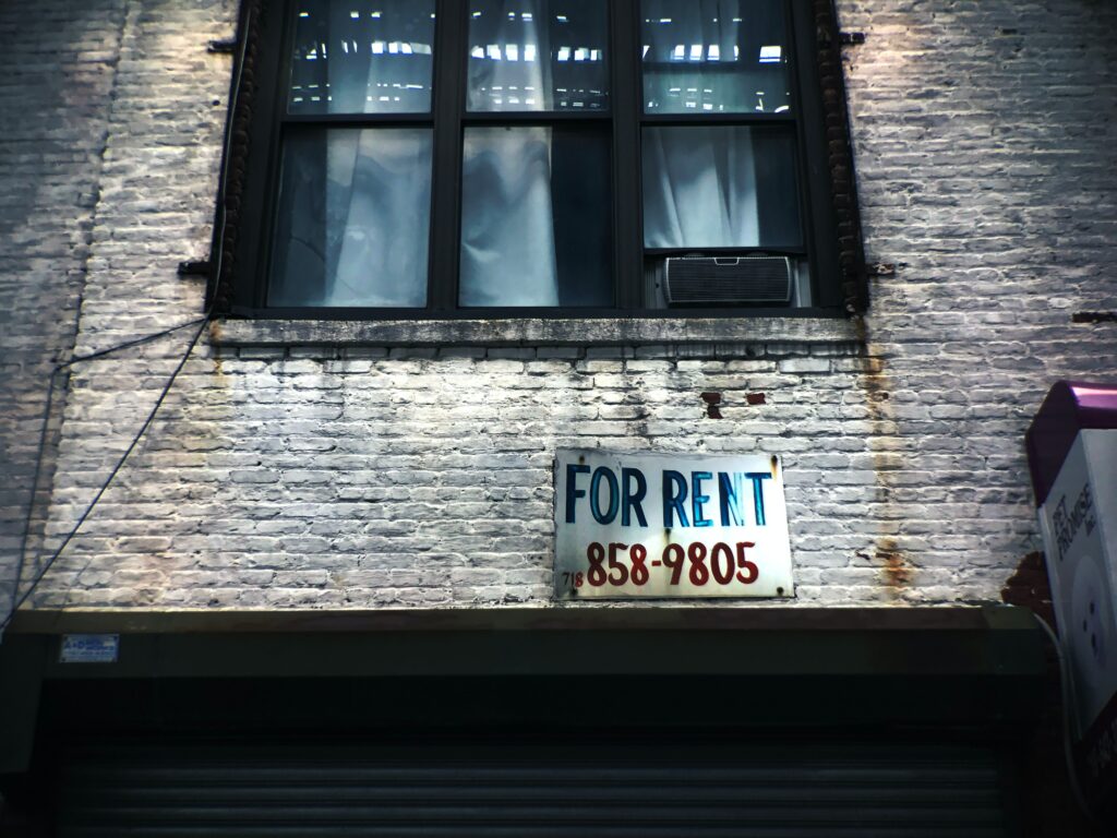 A For Rent sign below a window on a weathered building.