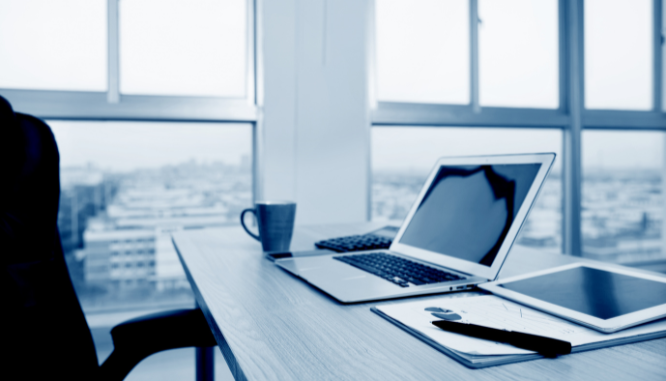 A desk with a laptop on it by some windows overlooking a city.