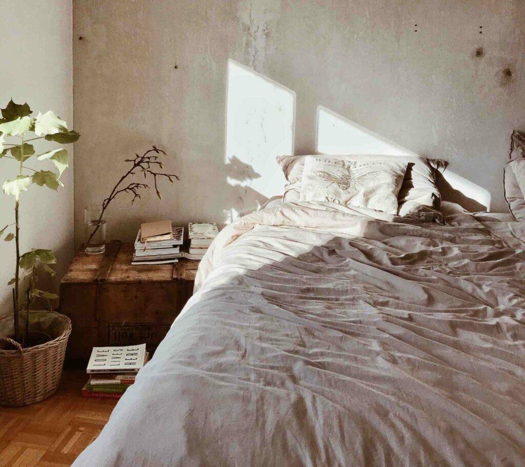 A bed in a guest room with morning light creeping in through a window.